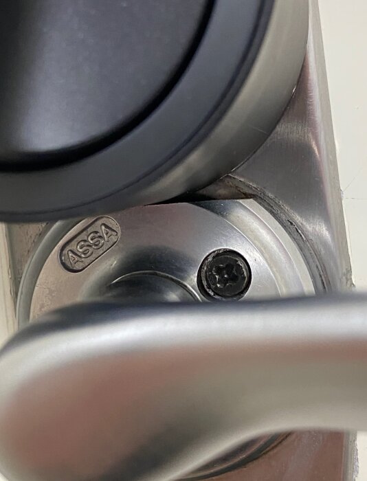 Närbild av ett ASSA 2002 lås där låsvredet är monterat upp och ner i en dörr, med borrskruv och en kapad bricka runt handtaget.