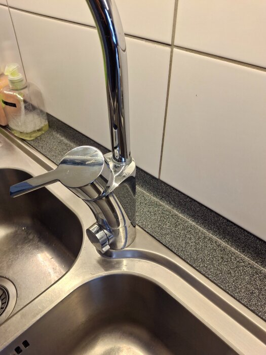 Köksblandare i rostfritt stål vid diskho, omgiven av kaklade väggar och ett handtvål vid sidan om diskbänken.