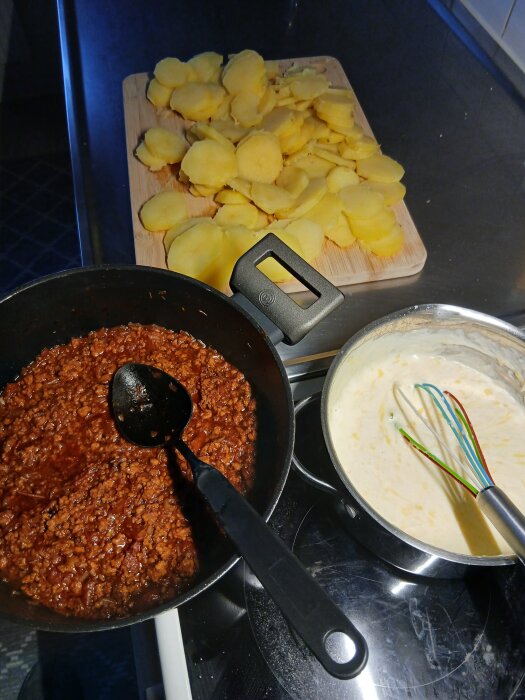 Potatisskivor, köttfärssås och vit sås redo för att lagas ihop till Skånsk moussaka i ett kök.