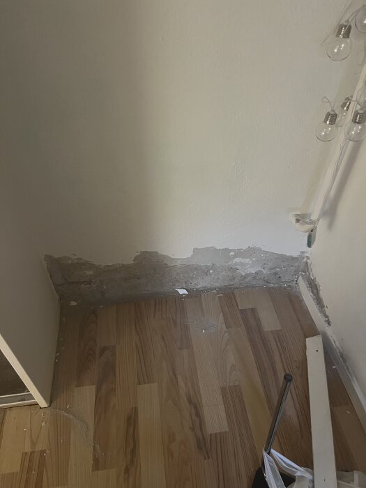 En källarvägg med avskalad puts och missfärgningar vid golvet. Trägolv är tydligt synligt och vissa verktyg ligger i hörnet.