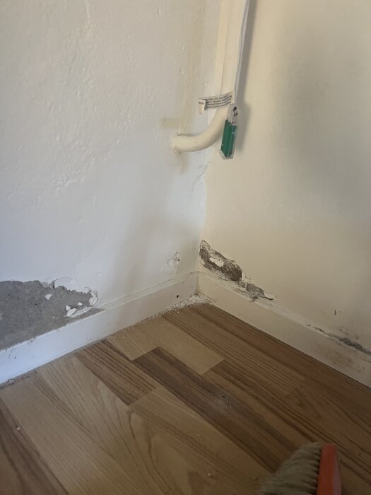 Vattenskada på en källarvägg vid ett trälaminatgolv, med synlig puts som flagnar och rör i hörnet, visar fuktproblem och behov av reparation.