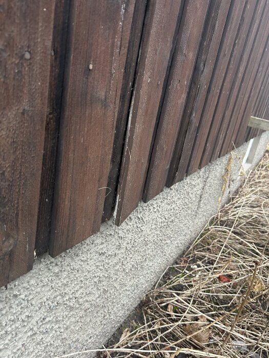 Träpaneler på en vägg med synligt glapp mellan paneler och vägg, nära marken med torr vegetation framför.