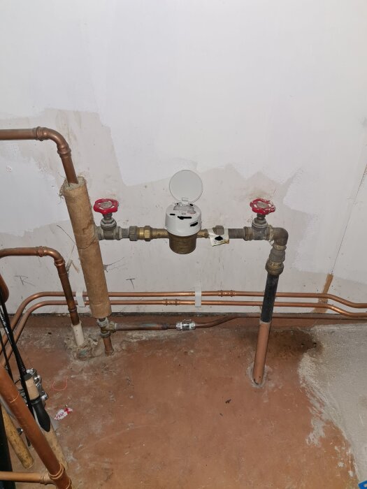 Vattenmätare med rörinstallation i källare, inklusive ventiler och kopplingar, mot en vit vägg och betonggolv.
