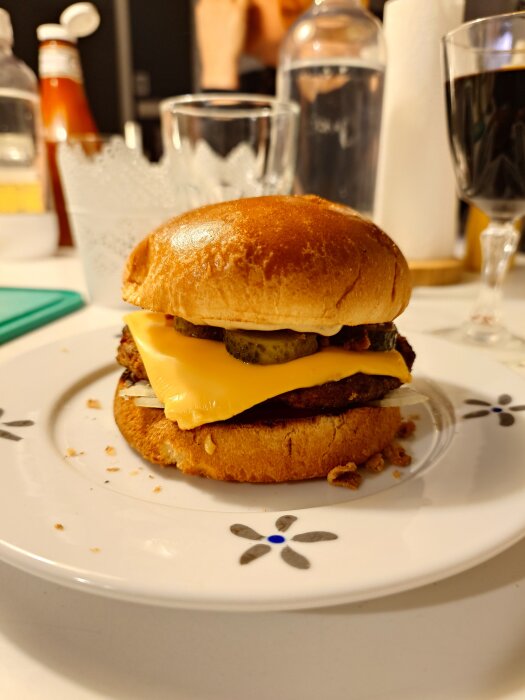 En närbild av en hamburgare med ost och pickles på en tallrik, omgiven av glas och flaskor på ett dukat bord.