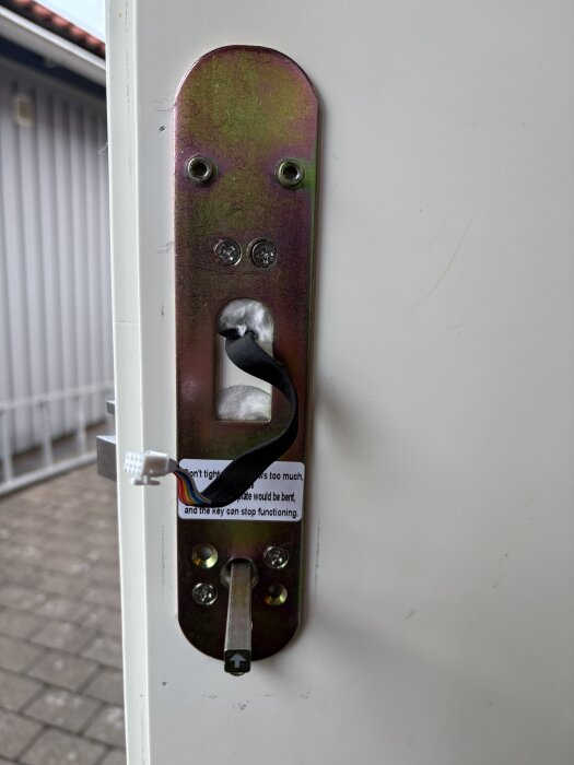 Inre mekanismen av ett lås installerat på en vit dörr med synliga skruvar och en kabel kopplad innanför ett hål, i en utomhusmiljö.