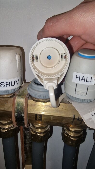 Värmefördelare med ventiler märkta "Sovrum" och "Hall". En hand håller i en av ventilerna, kopplad till rören under.