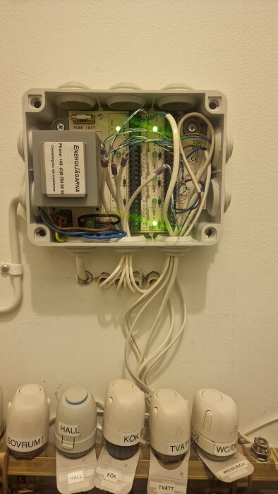 Kopplingsbox med kablar och elektronik, under finns ventiler märkta för olika rum: sovrum, hall, kök, tvätt och WC/dusch.