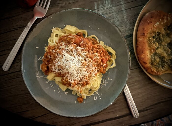 Färsk fettuccine med köttfärssås och riven Parmigiano-Reggiano, serverad med vitlöksbröd och en gaffel på ett träbord.