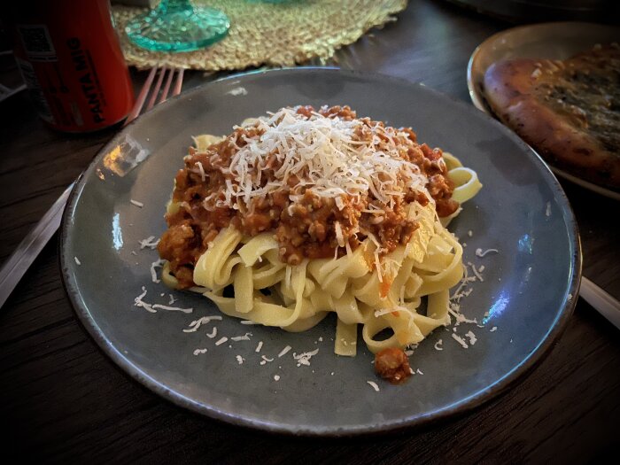 Färsk fettuccine med bolognese och riven Parmigiano-Reggiano serverad på en tallrik, med vitlöksbröd vid sidan och en drickaburk i bakgrunden.