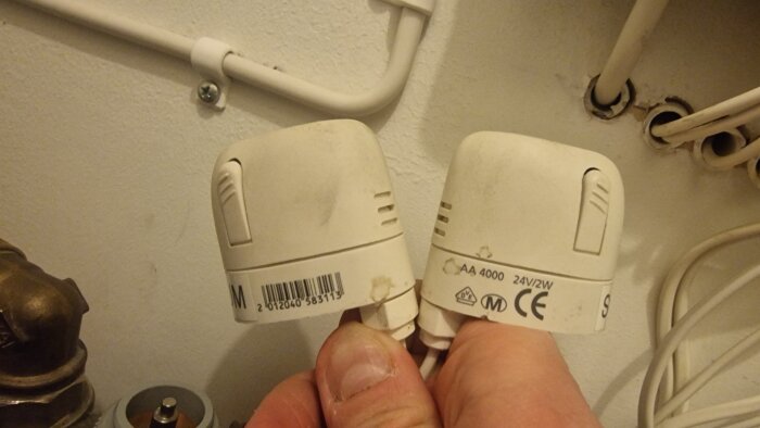 Två radiatorventilstyrningar hålls i handen, monterade vid väggen med rör och kablar i bakgrunden.
