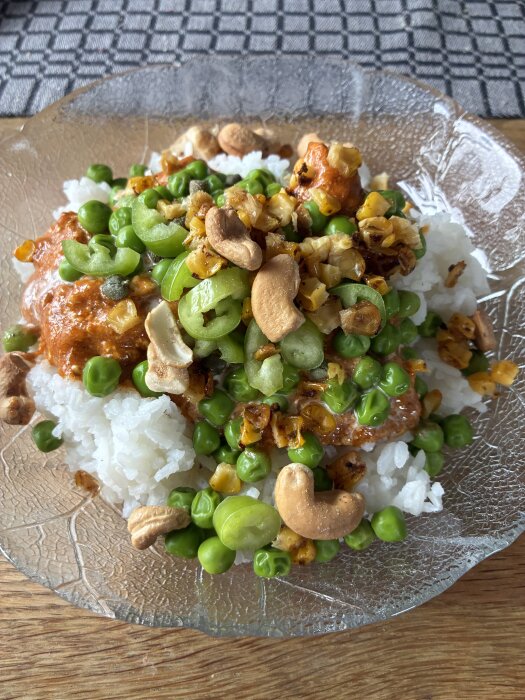 Risrätt toppad med ärtor, cashewnötter, grillad lök och sås i en glasassiett.