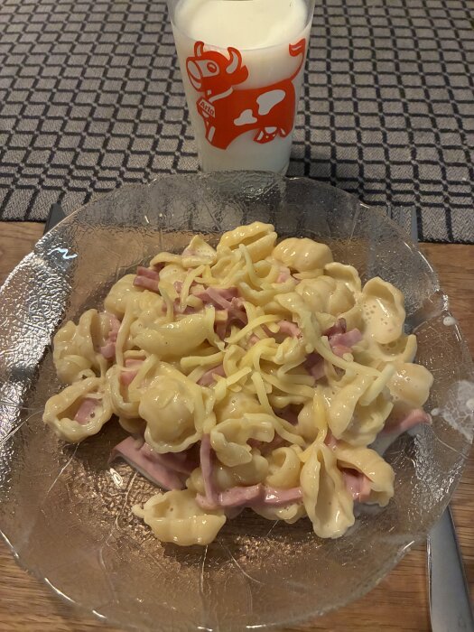 Pasta med ost och skinka på tallrik, med ett glas mjölk i bakgrunden på bord med mönstrad duk.