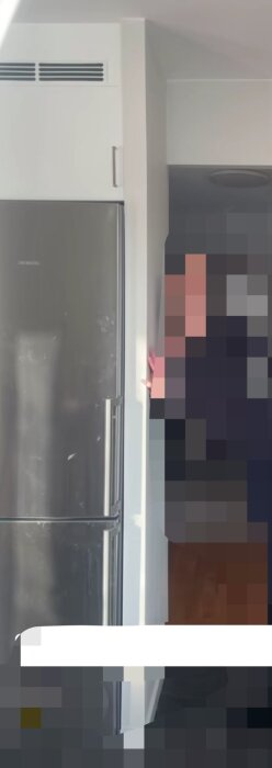 Kylskåp i rostfritt stål vid sidan av en vit vägg med delvis suddig person i bakgrunden.