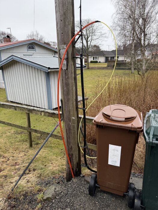 Orange och gul fiberkabel hänger på en trästolpe bredvid en brun soptunna och en grön soptunna i en trädgårdsmiljö.
