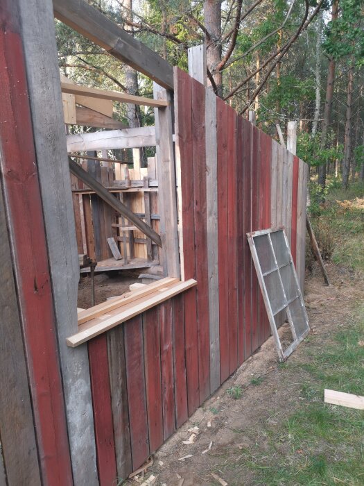 Gamla träbrädor utgör en delvis byggd vägg vid ett garageprojekt, med fönsterkarm lutad mot väggen i en skogsmiljö.