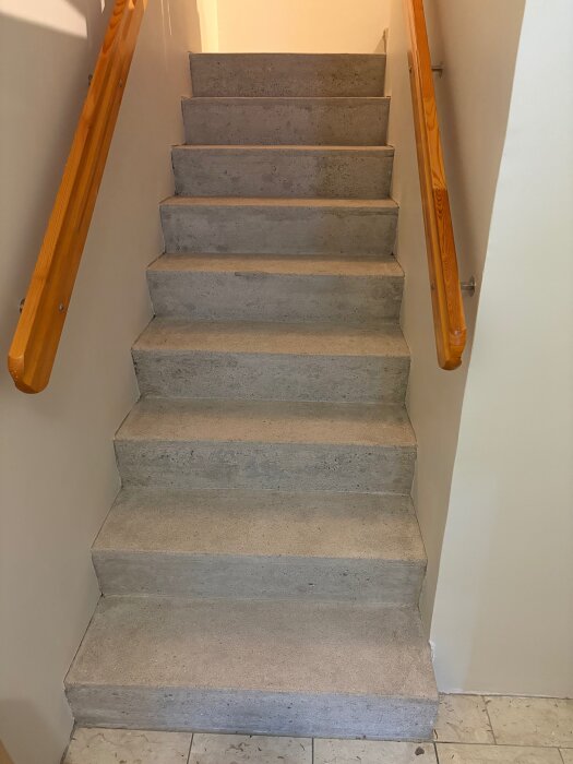 Betongtrappa inomhus utan matta, med trähandledare på vita väggar, upp till vardagsrum.