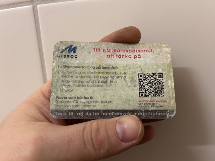 Person håller i ett kort från Midroc med instruktioner för sjukvårdspersonal angående läkarmätningar och prover, inklusive en QR-kod.