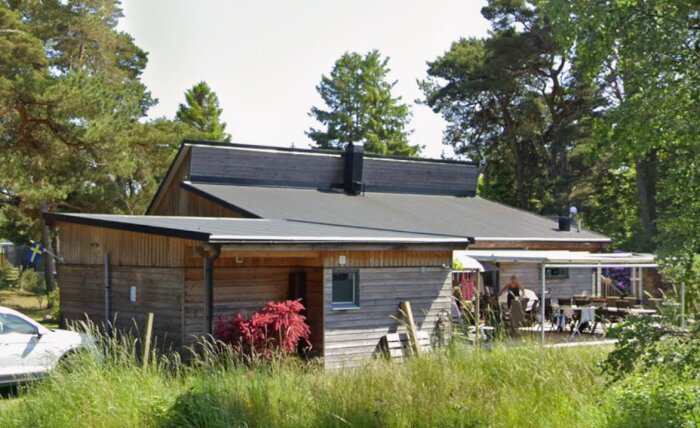 En lutad skorsten på ett trähus med platt tak, omgiven av träd och en flagga.