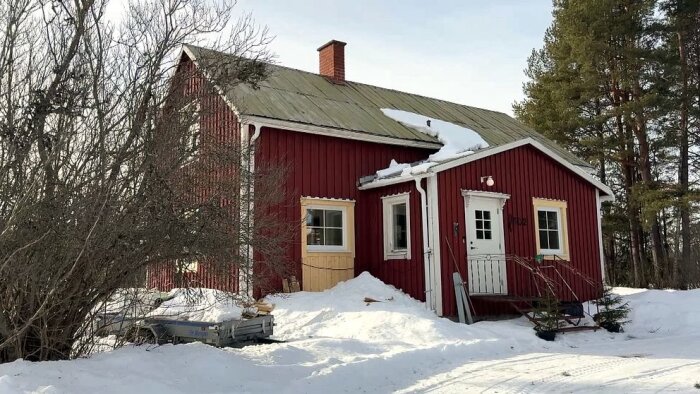 Rött trähus med snö på taket och omgivningen, ute på landsbygd med barrträd i bakgrunden, fotograferat i dagsljus.