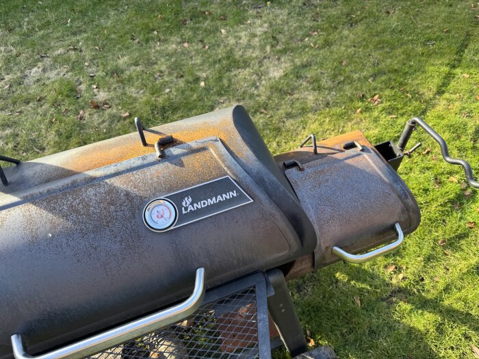 Offset-smokergrill på gräsmatta, delvis rostig, med "Landmann"-logotyp och temperaturmätare, redo för vård och inoljning.