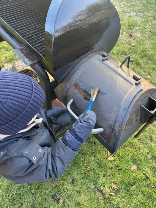 Person oljar in en offset smoker med en pensel i en trädgård.