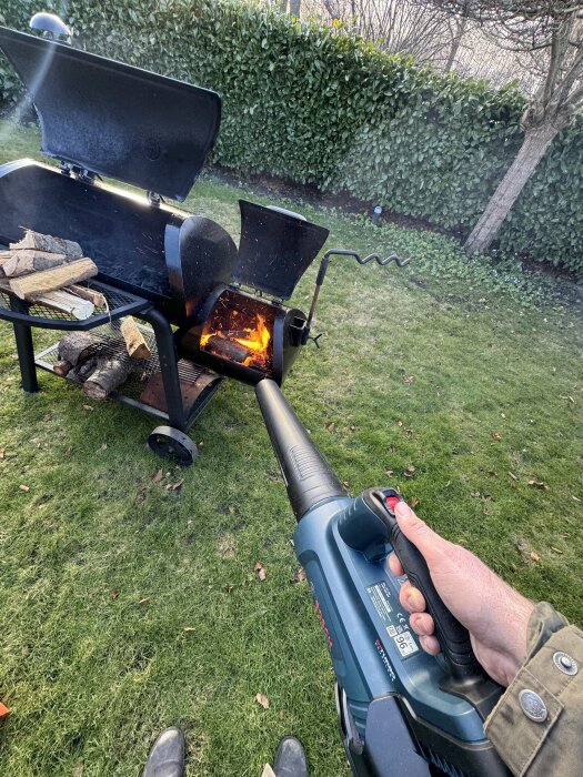 Person använder lövblås för att få fart på elden i en offset smoker grill. Brinnande ved i grillen, gräsmatta och häck i bakgrunden.