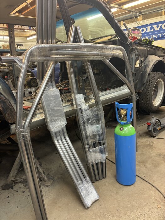Stålram och stålrör i ett garage, intill en blå gasflaska, en del av ett bilbyggprojekt med en bil i bakgrunden.