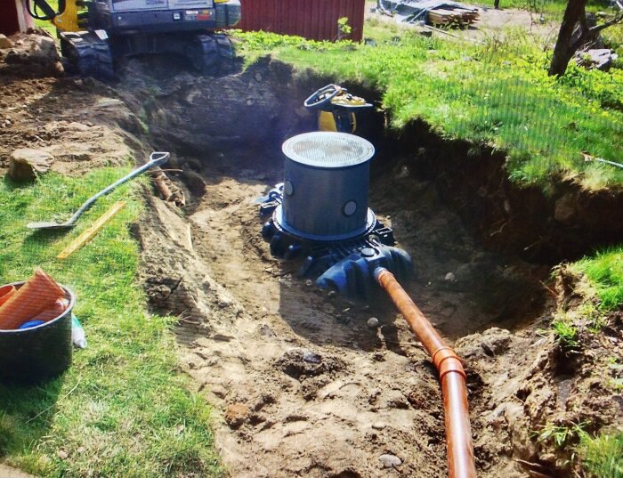 Grävd grop med en avloppspumpbrunn installerad, omgivna av jord och med en orange rörledning ansluten.