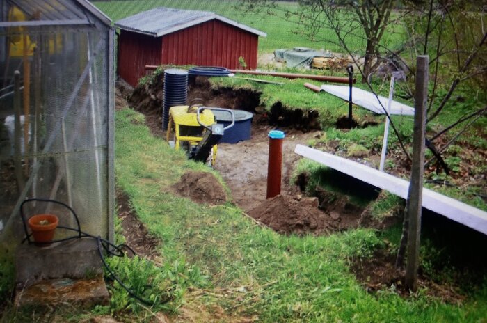 Grävplats för avloppssystem med brunnar och rör, en pump och redskap syns i en trädgård med växthus och gräsmatta.
