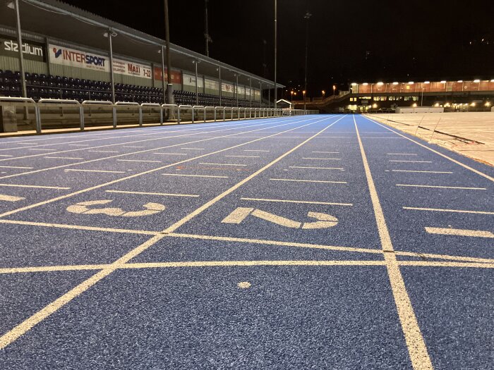 Blå löparbana med vita markeringar och startnummer i ett upplyst stadionområde på natten.