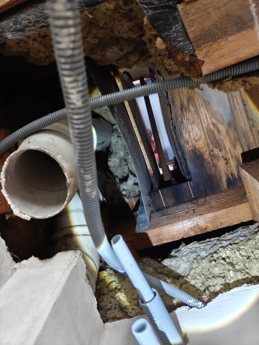 Självdrags-ventilationschakt på vind med masonit mot plåt, isoleringsmaterial saknas, rör och elkablar synliga.
