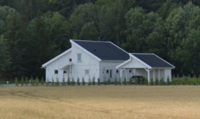 En vit villa med svart tak i ett landskap omgiven av höga buskar, en carport syns till höger framför garaget.