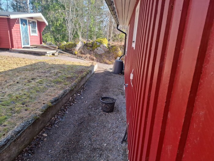 Röd husfasad med gammal stödmur bredvid grusgång. I bakgrunden syns träd och stenar. En svart plastbehållare står på marken.