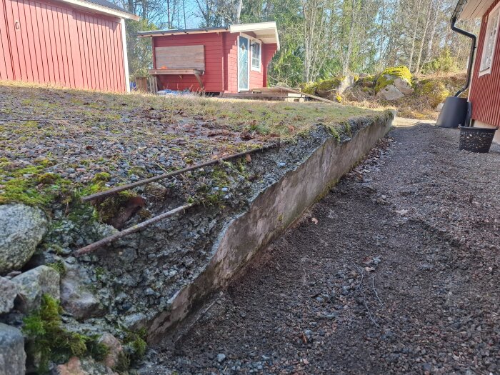 Gammal och delvis förstörd stödmur bakom ett hus, med synlig armering och mossbeläggning. Omgiven av röda byggnader och stenar i bakgrunden.