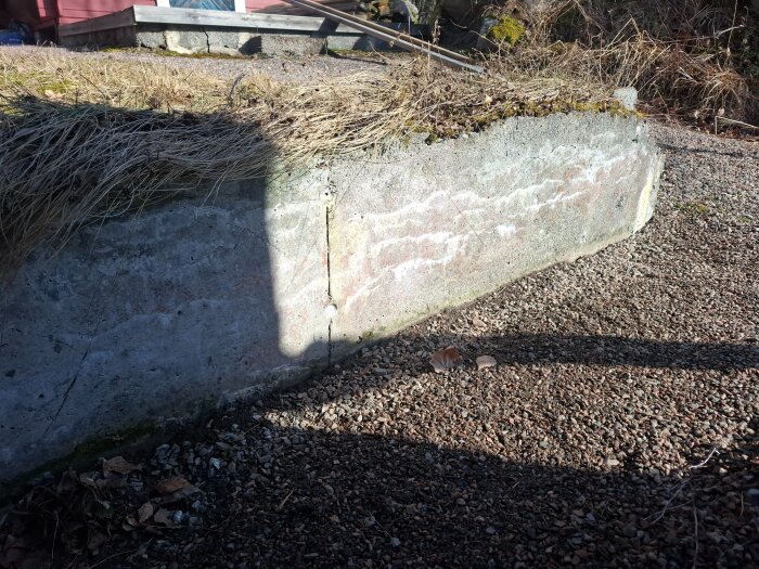 En gammal stödmur med skador och sprickor, omgiven av torrt gräs och grus. Murens skick tyder på behov av reparation eller ersättning.