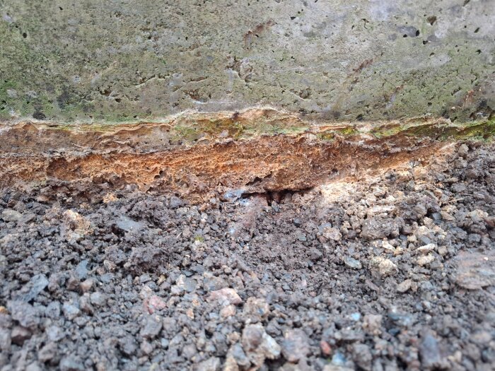 Närbild på en gammal stödmur med tydliga sprickor och erosion vid basen, omgiven av jord och grus.