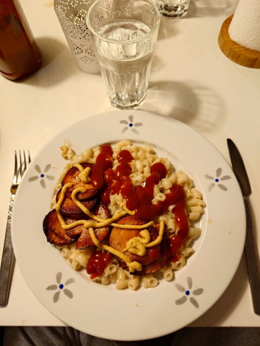 Tallrik med pasta, stekta korvskivor, ketchup och senap. Vattenglas och en servetthållare i bakgrunden.
