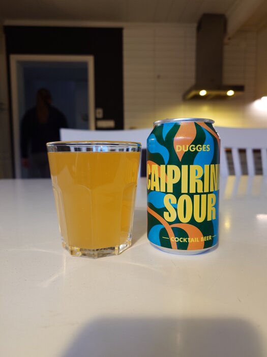 Burk med Dugges Caipirinha Sour cocktail beer bredvid ett glas med gul dryck på ett vitt bord i ett kök.