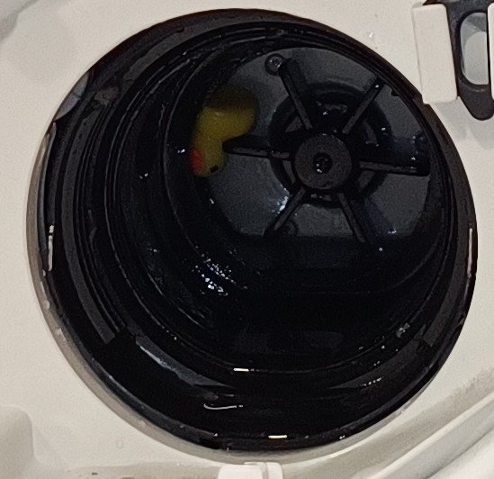 Gul anka inuti tvättmaskinens filteröppning med svart inre och ventilformad insats.