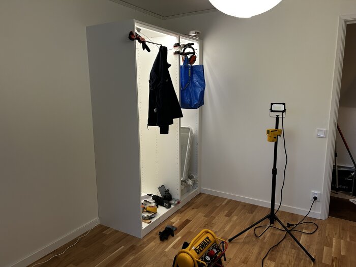 Klädhängare i garderob med kläder och IKEA-påse, vägglampa på stativ och verktyg på nytt Kährs Ek Lecco-golv.