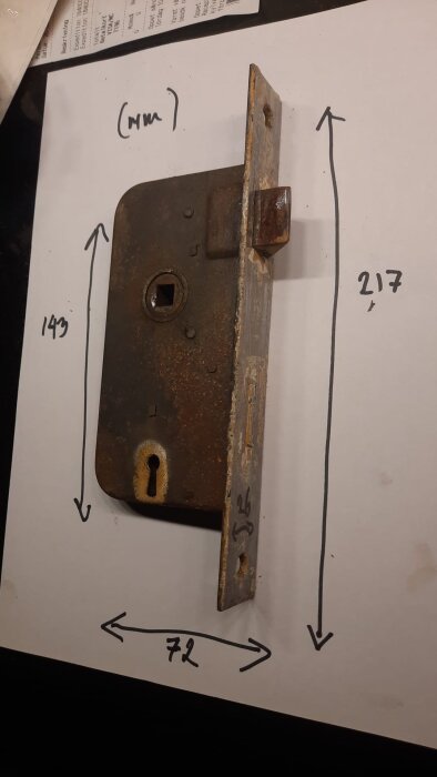 Rostigt gammalt dörrlås av metall med mått markerade på papper: 217 mm hög, 72 mm bred och 143 mm lång.