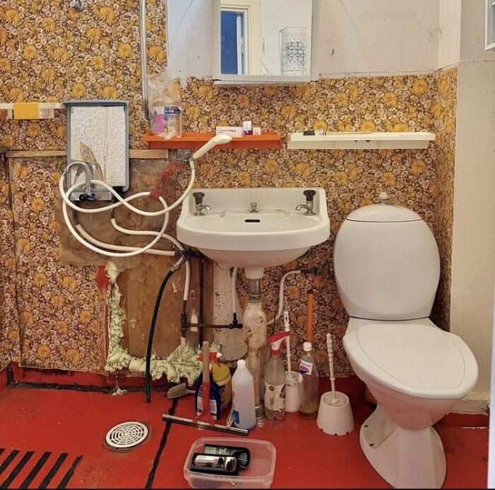 Badrumsrenovering pågår med ett handfat, toalett och blommigt mönstrade väggar, samt olika rengöringsmedel på golvet.