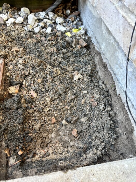 Grävd grop vid husgrund visar lager av jord, sten och krossmaterial. Synligt fiberduk delvis under krossen vid sidan av en husvägg.