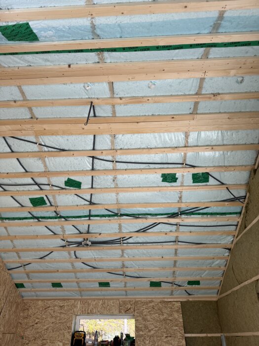 Tak med nyinstallerad lösullsisolering och exponerade träbalkar, sett inifrån ett rum under renovering, med kablar dragna ovanför isoleringen.