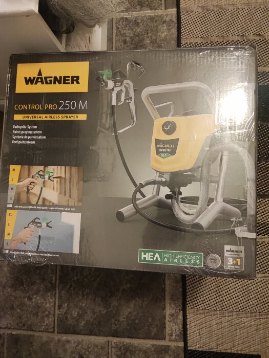 Förpackning av sprutpistol Wagner Control Pro 250 M, universal airless sprayer, på ett kaklat golv med matta.
