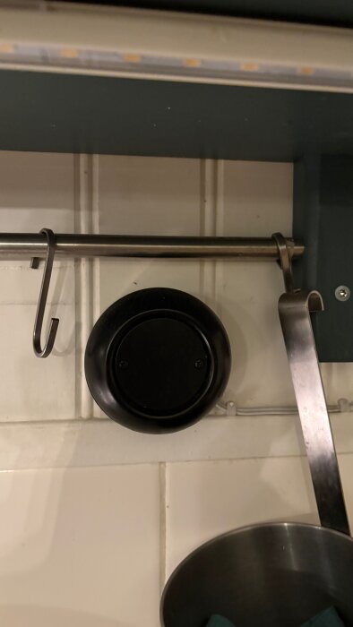 Svart rund enhet på väggen under kökshylla med metallstång och krok, ospecifierad funktion, med LED-list monterad ovanför.