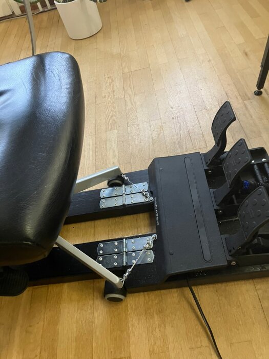 Stol för bilspel monterad intill pedaler med två träskruvar på trägolv.