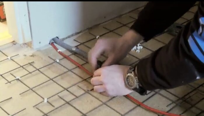 En person installerar röda och grå rör för golvvärme under klinkergolv, med väggöppning i bakgrunden.