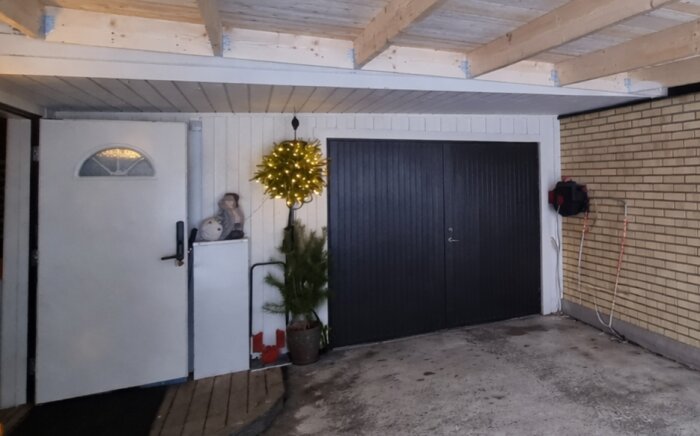Garage med två svarta slagportar och en sidodörr, dekorerat med granris och en ljusslinga över dörren.