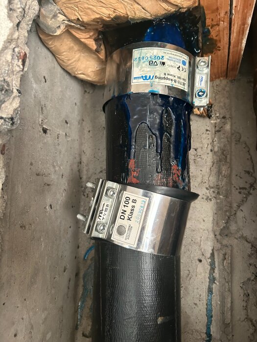 Svart avloppsrör med stålfästen och blå tätning, omgiven av trä och betongvägg. Märkning med texten "DN 100 Klass B".